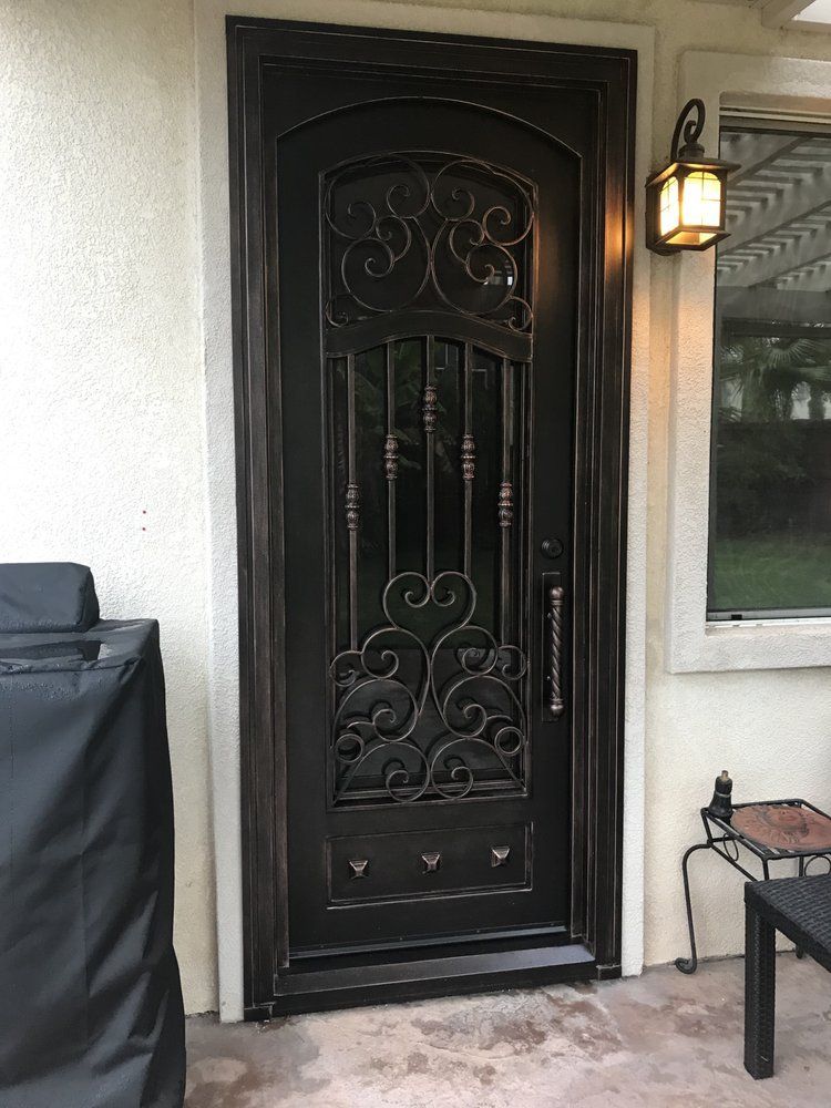 Ornate black wrought iron door with glass, set in a light beige frame, with a wall lantern beside it.