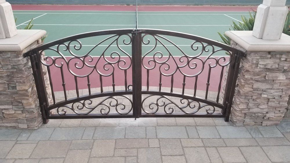 Ornate wrought iron double gate with a curved top, set between stone columns, opening to a tennis court.