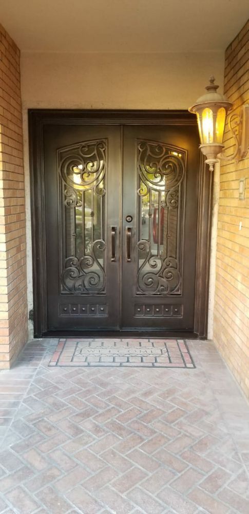 Ornate dark double doors with decorative glass panels and a welcome mat on a brick patio.